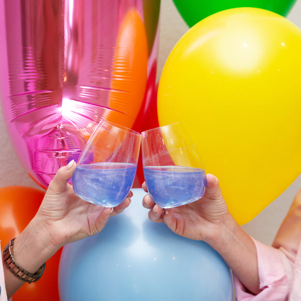 Two stemless glasses with Sugar Mama Shimmer Little Lilac edible glitter for drinks doing a "cheers" in front of birthday balloons.
