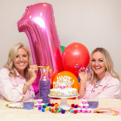 Two blonde women celebrating Sugar Mama Shimmer's birthday with cake, balloons, party horns, and Sugar Mama Shimmer Little Lilac edible glitter for drinks in stemless wine glasses.