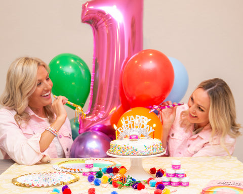 Two blonde women celebrating Sugar Mama Shimmer's first birthday with balloons, party horns, cake and Sugar Mama Shimmer Little Lilac edible glitter for drinks in wine glasses.