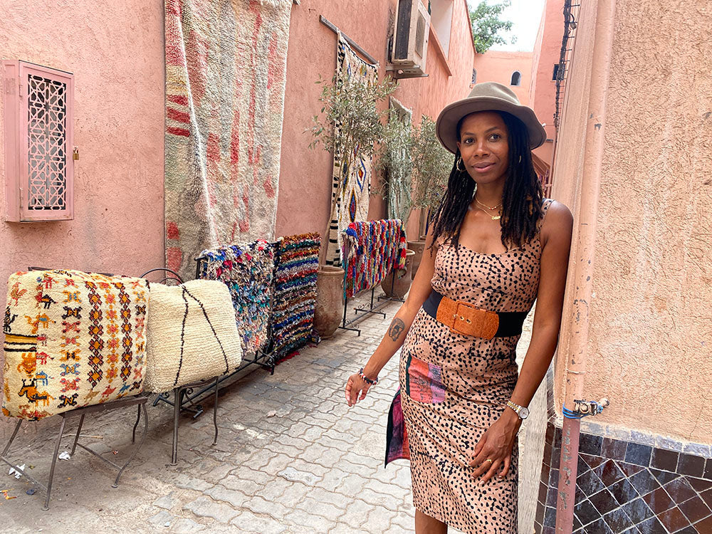 Nasozi is a Medina alleyway standing in front of Berber and Beni Ourain Moroccan rugs and poufs. The wall she is leaning on is made of traditional ceramic Moroccan tiles.