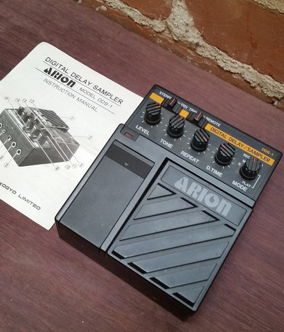 black rectangular effects pedal with five knobs. white text. also pictured a small black & white booklet to the left of the pedal.