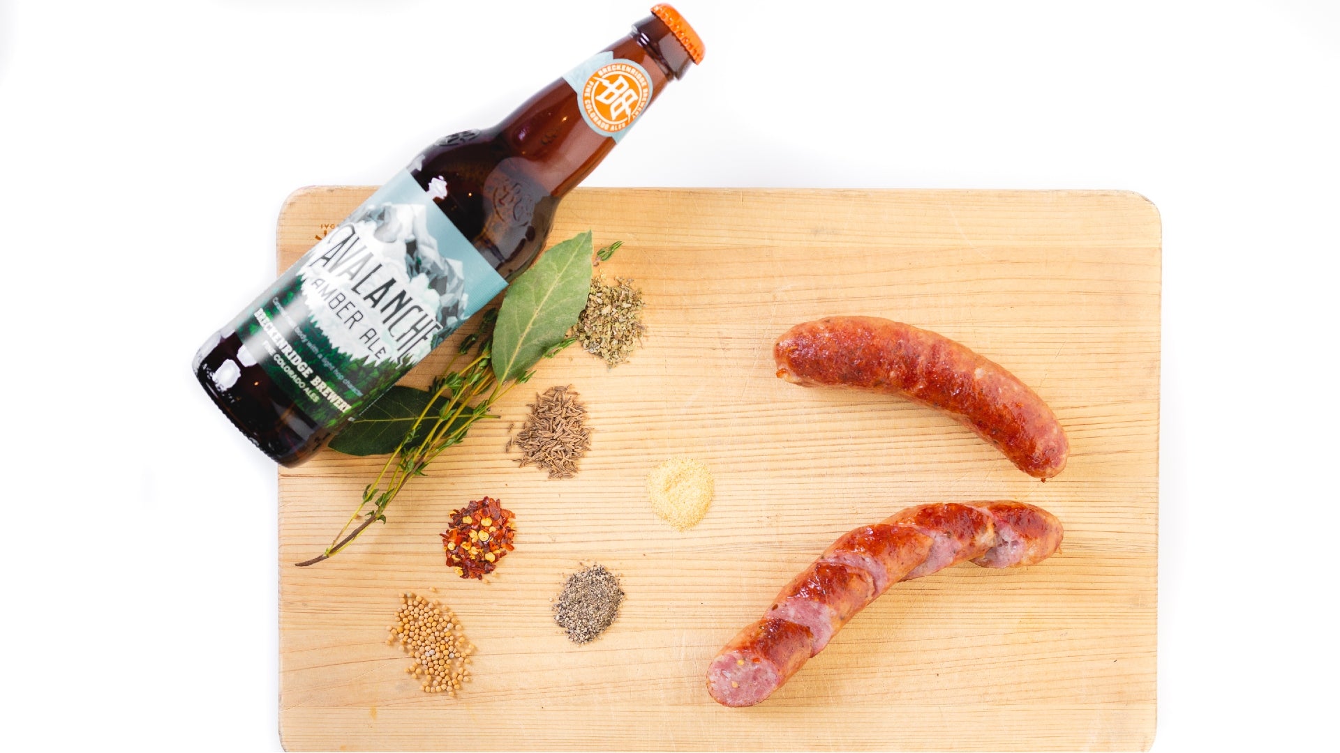 A picture shot from above of an Avalanche smoked cheddar bratwurst on a cutting board with herbs, spices and the Breckenridge Avalanche beer.