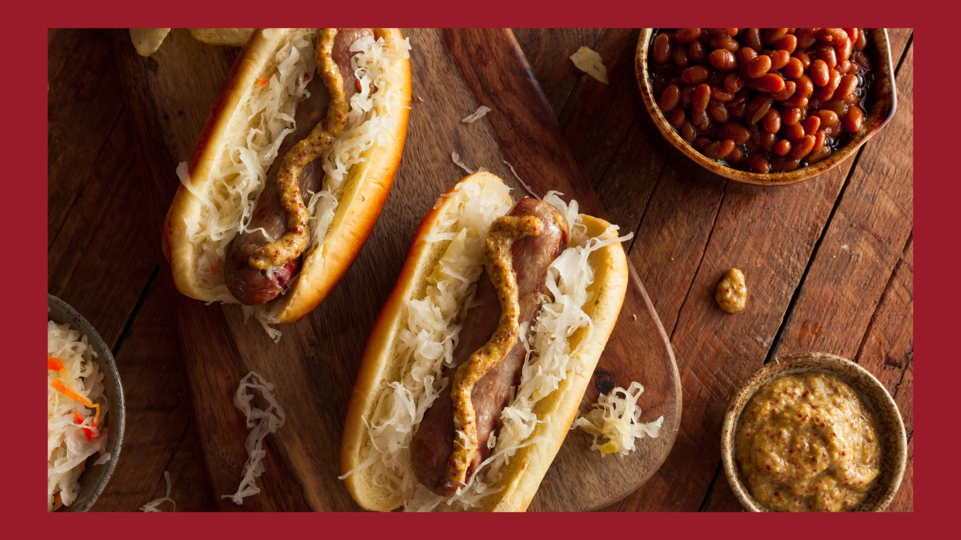 Bratwursts in buns and topped with sauerkraut, onions, and brown mustard on a wooden cutting board. 