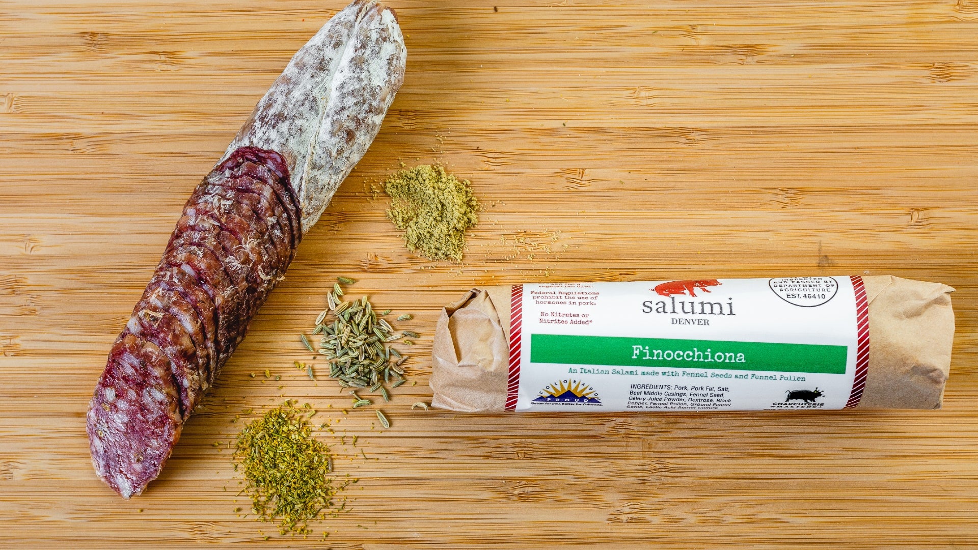 Two pieces of Finnochiana salami, one in packaging and one cut into pieces, on a cutting board with spices in between them.