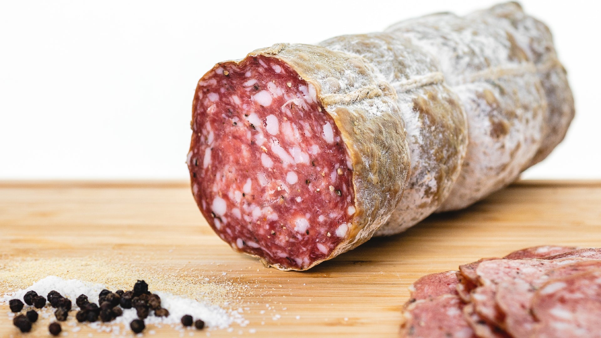 The cross section of a chub of Genoa salami on a cutting board next to spices and with genoa slices in the foreground.