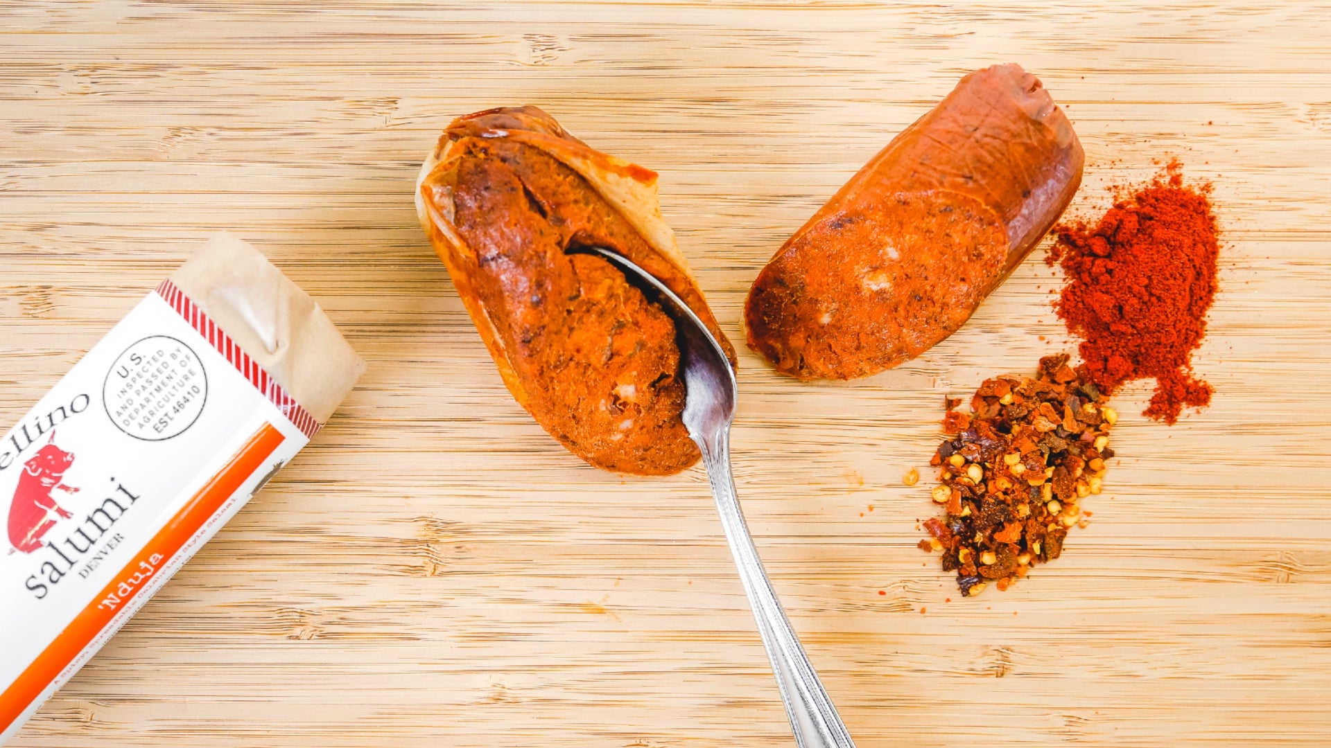 A picture shot from above of 'Nduja Salami on a cutting board. One chub of the salami is in it's packaging while the other chub has a spoon cutting into it to show that it's spreadable.