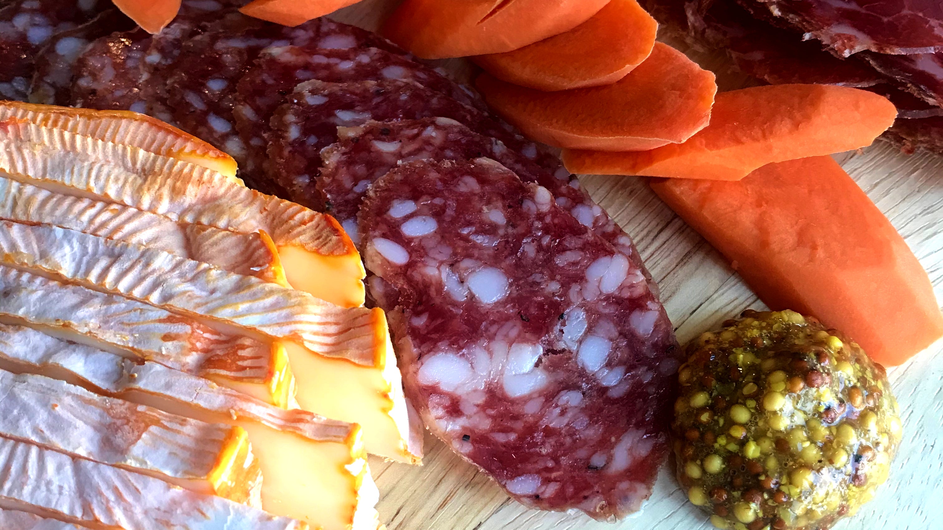 A close picture of il porcellino's rosette de lyon salami cut into slices on a cutting board. The salami is surrounded by pickled carrots, cheese and mustard.