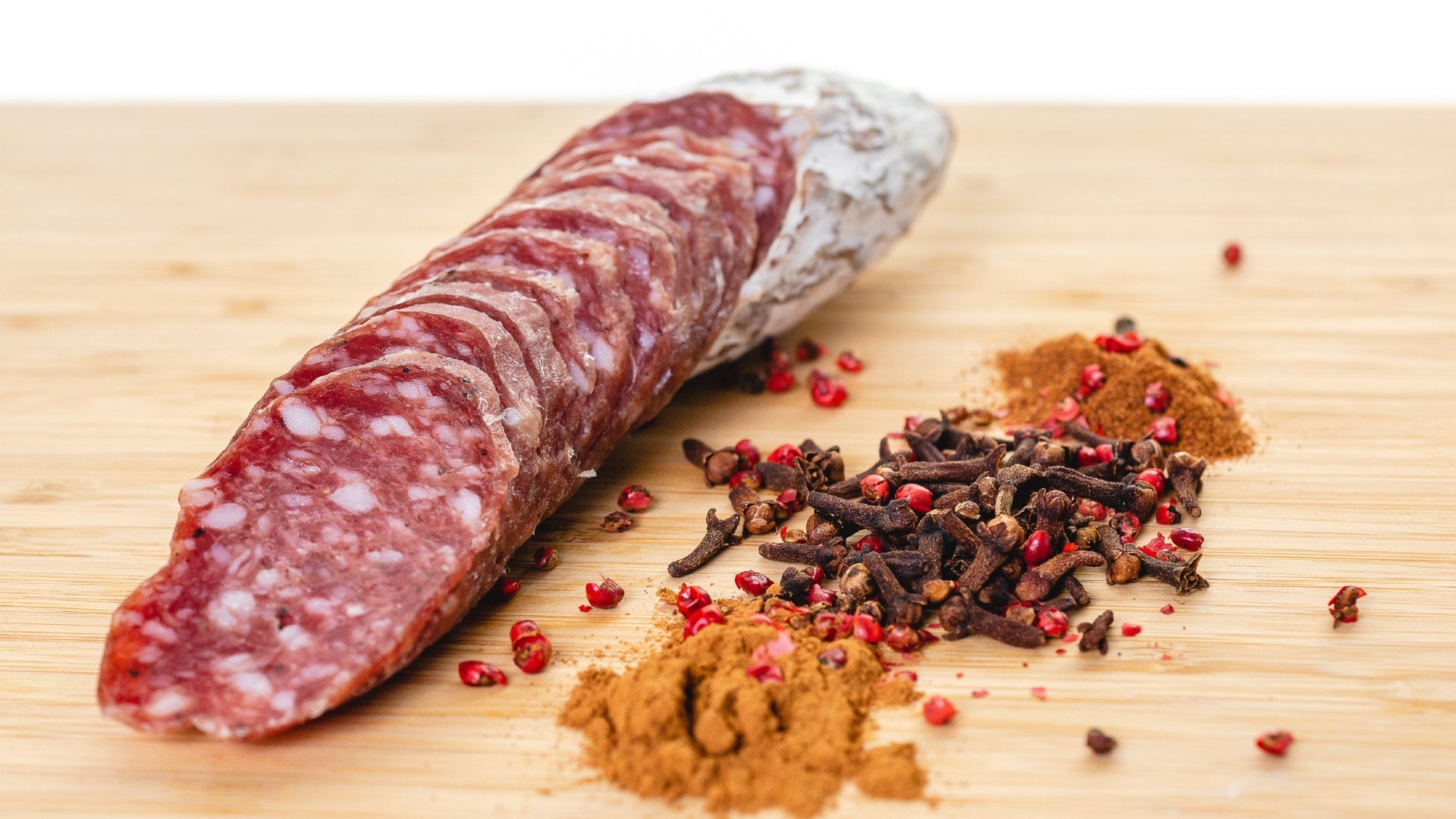 A close up of Rosette De Lyon salami cut into slices on a cutting board next to herbs and spices.