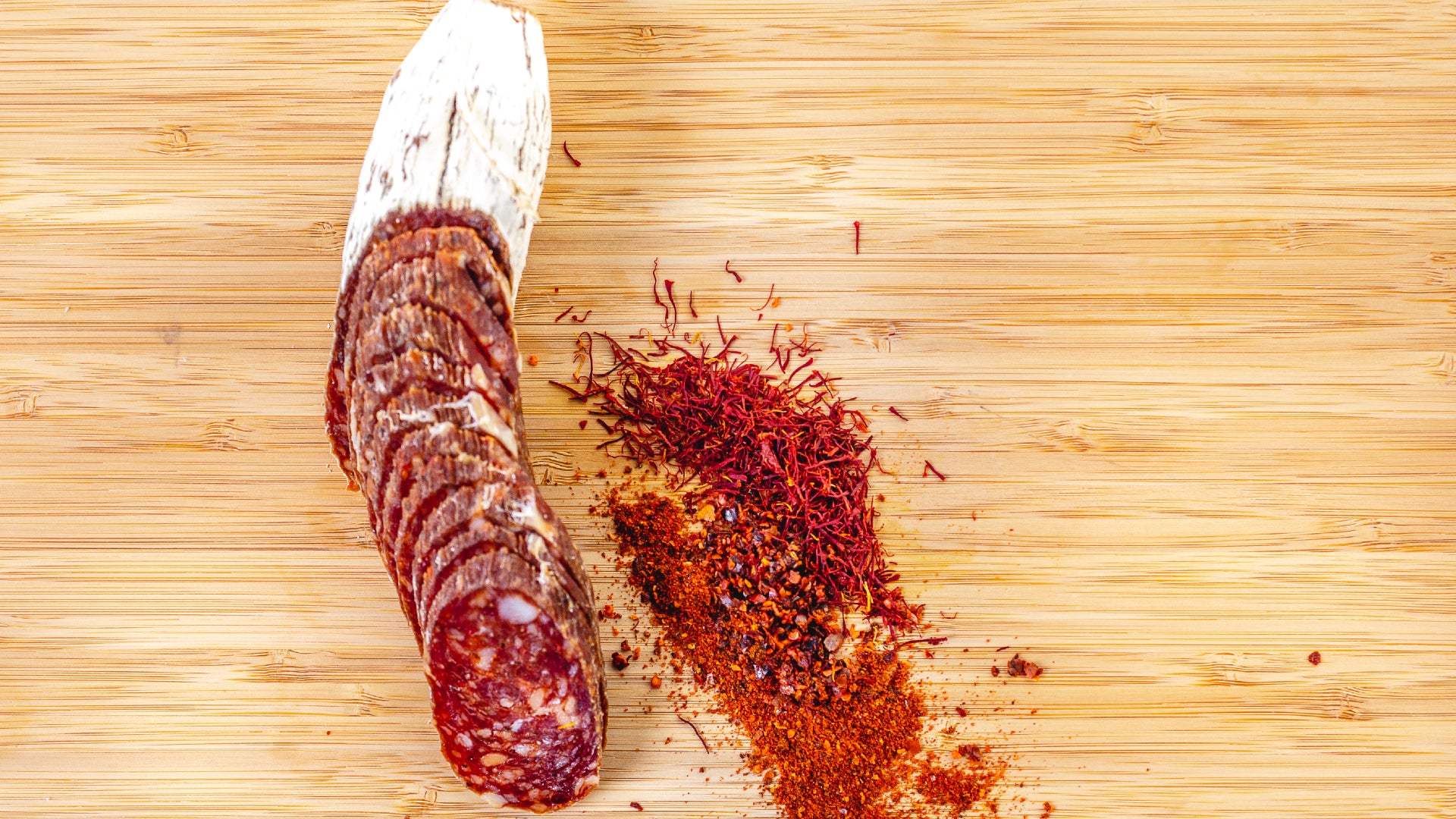 A picture shot from above of Saucisson Basque salami cut into slices on a cutting board next to herbs and spices.