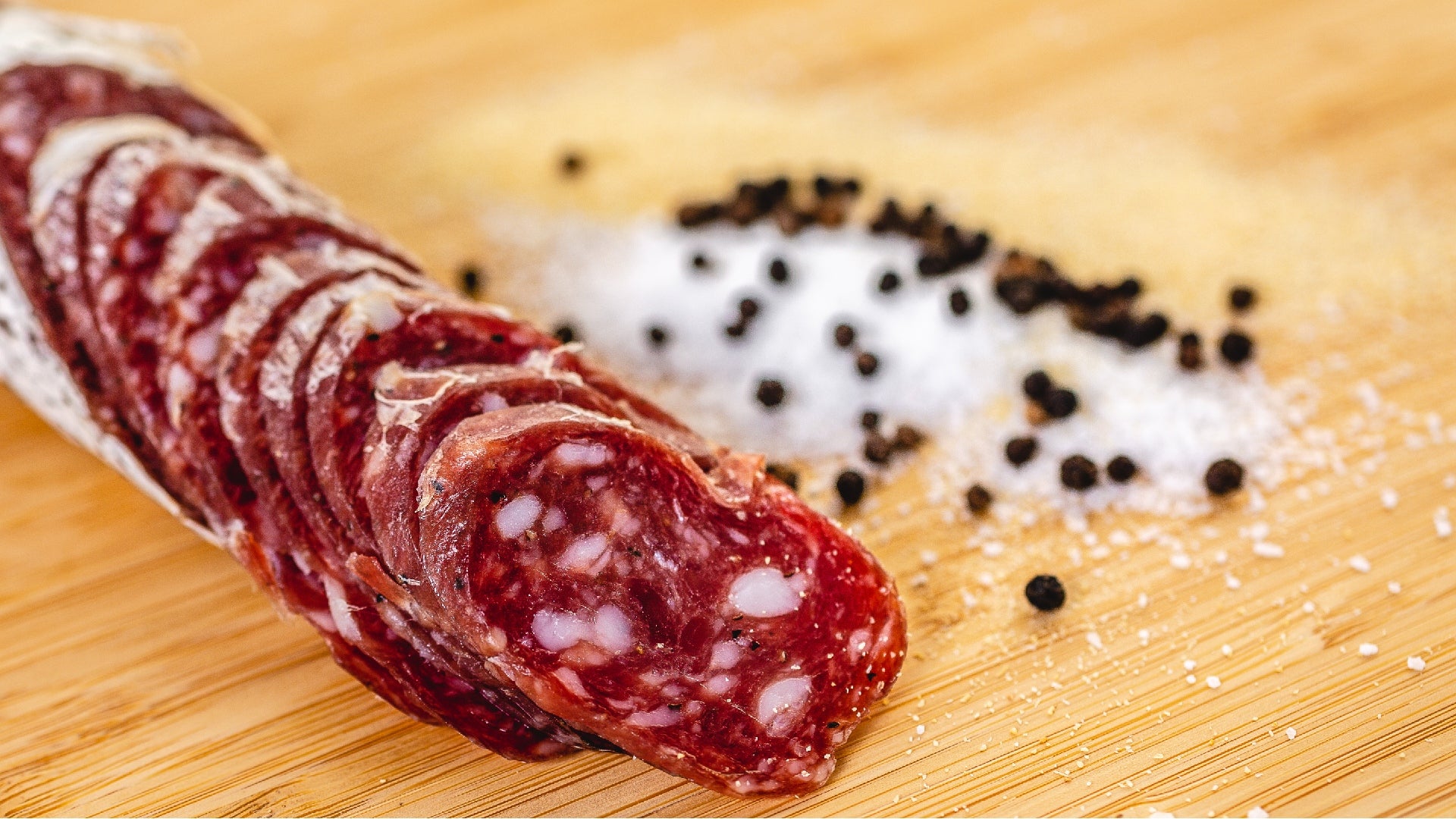 A close up of slices of Saucisson Sec salami on a cutting board with salt and pepper piled on the cutting board in the background.