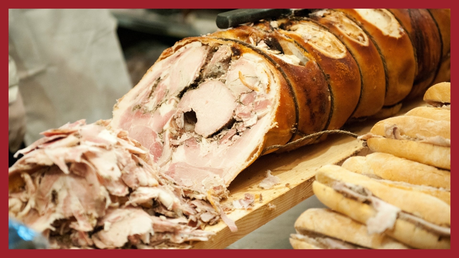 A whole porchetta being cut up into thin slices with porchetta sandwiches in the foreground.