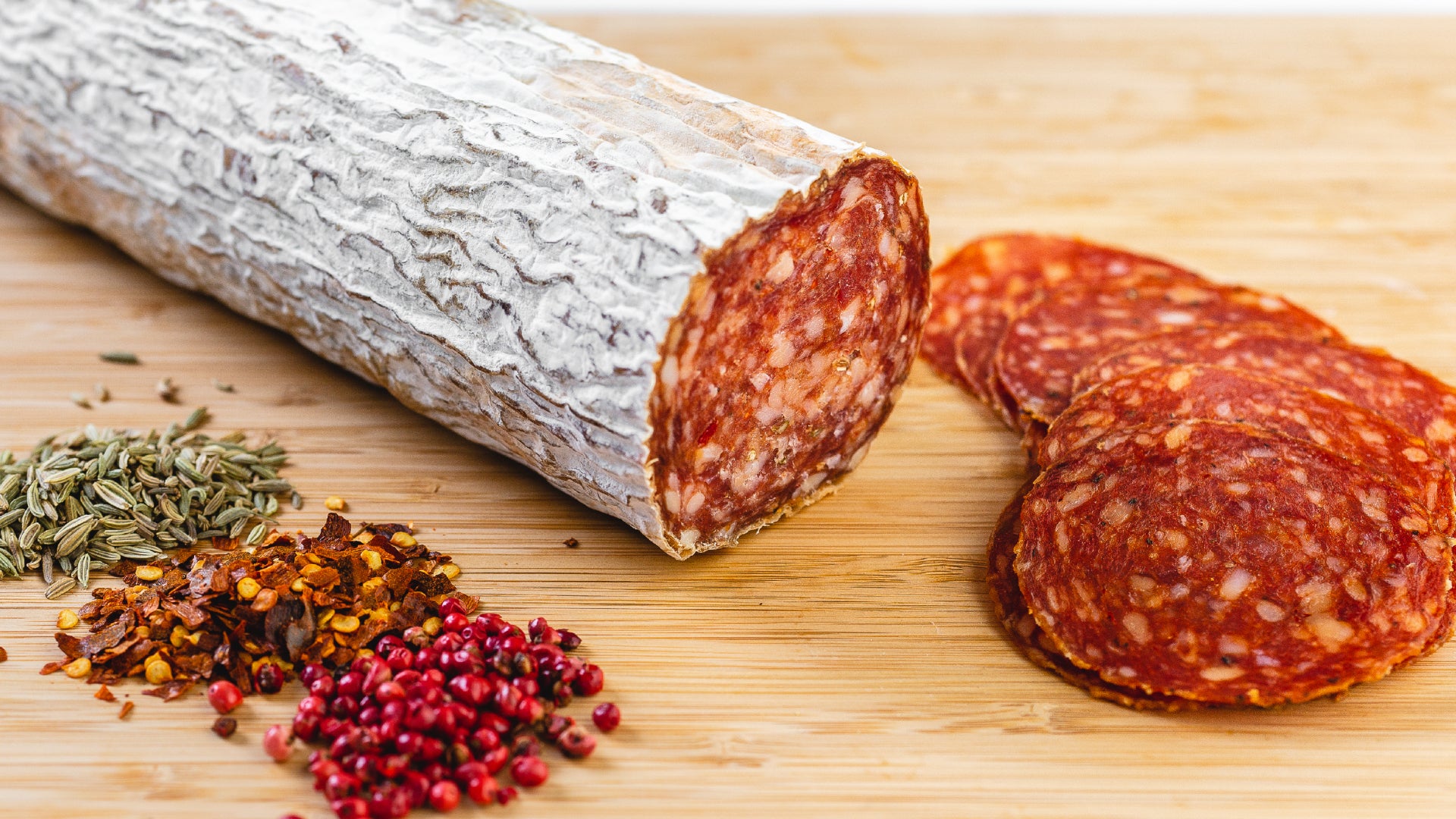 A large chub of Soppressata salami on a cutting board next to herbs and spices and with salami slices in the foreground.