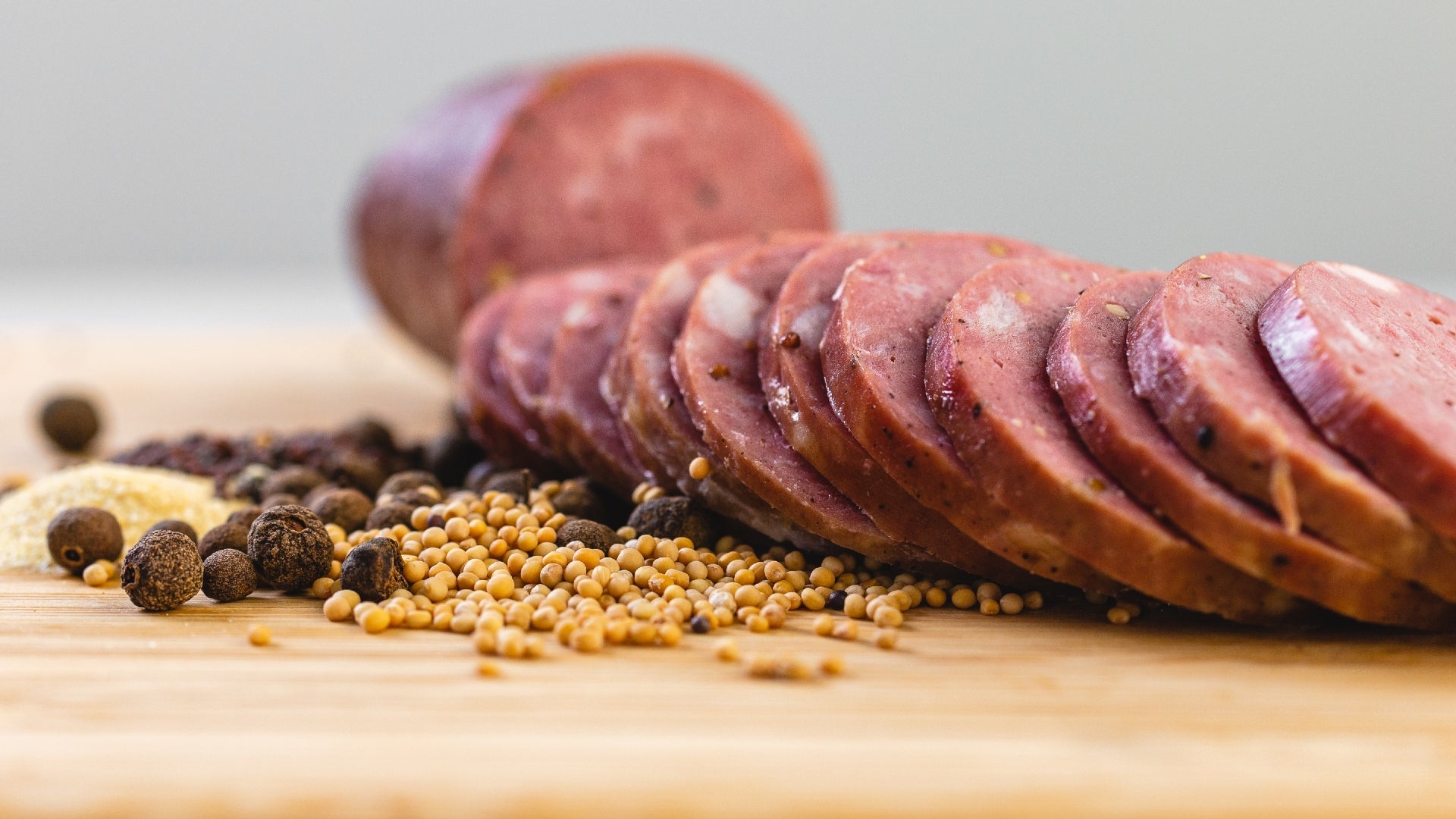 A close up of smoke summer sausage slices on a cutting board next to herbs and spices and with a whole chub of summer sausage in the background.