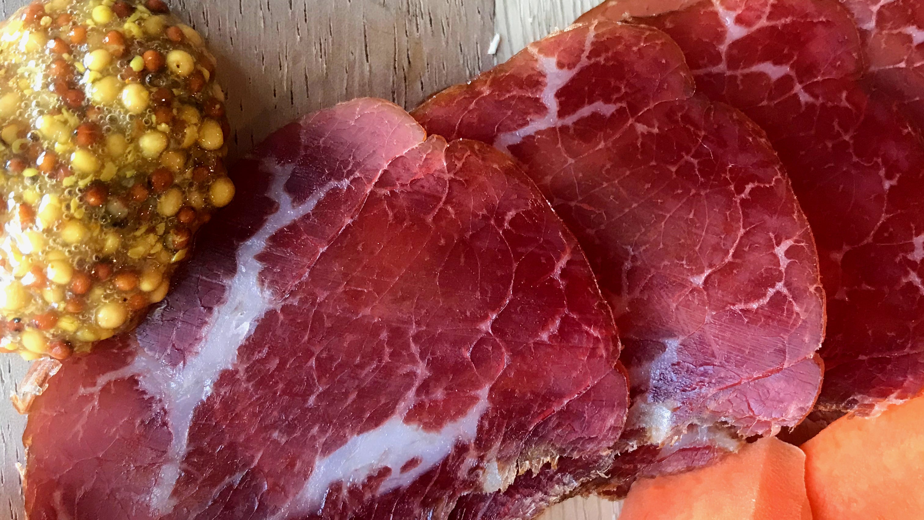 A picture of waygu beef bresaola shot up close and above. The bresaola is cut into slices and displayed on a charcuterie board next to mustard and carrots.