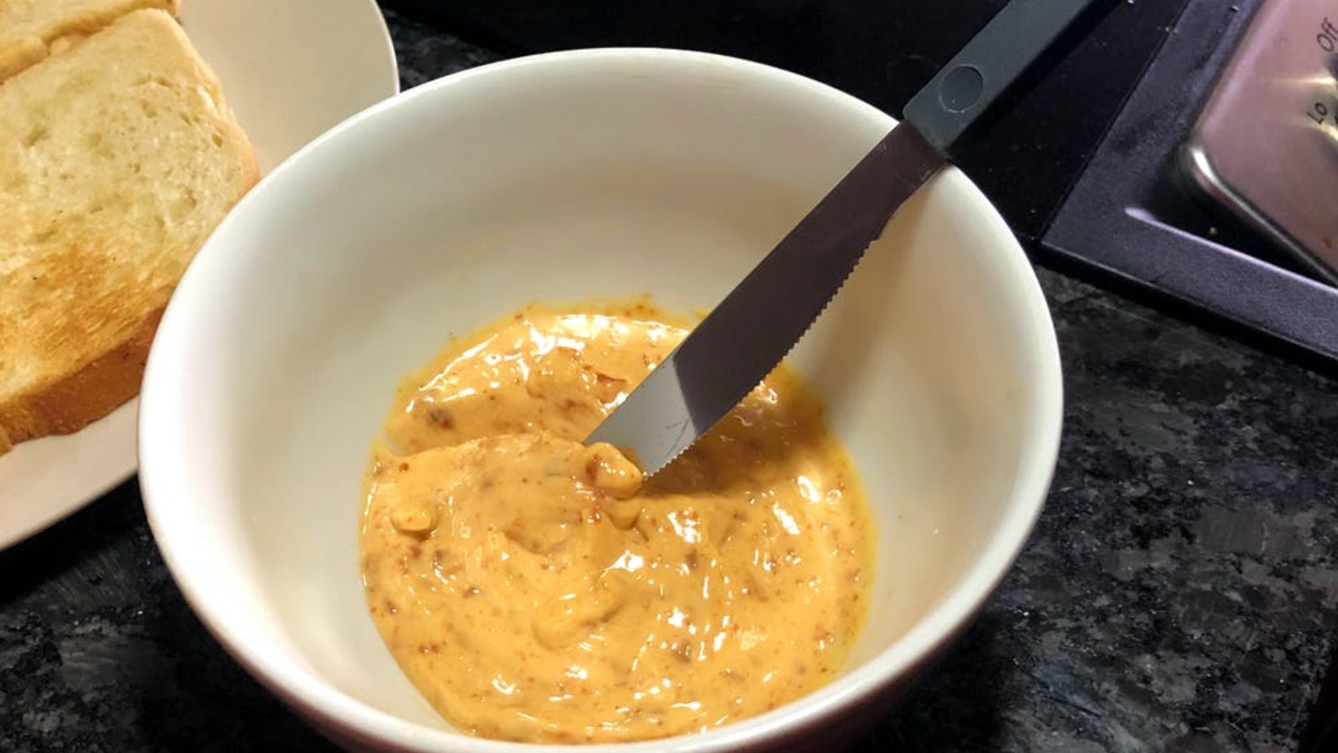 A bowl holding 'nduja salami mixed with mayonnaise with a butter knife in the bowl and toasted bread to the left of the bowl.