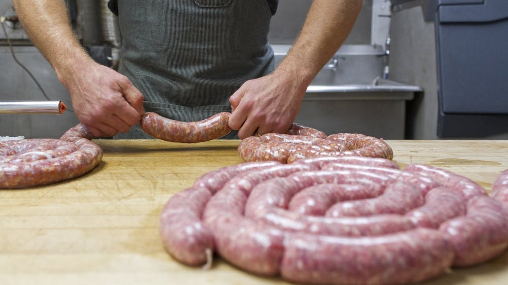 A picture showing someone tying off individual sausages from a large sausage link. Plus several rolls of linked sausage already prepared and resting on a butcher table.