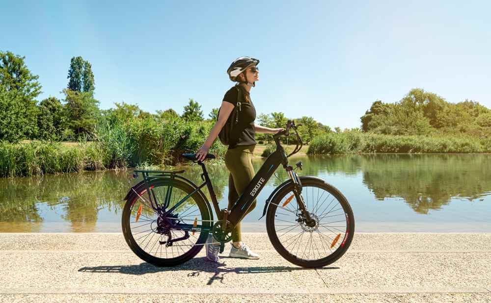 A woman wearing a helmet riding an electric hiking bike stands near a lake