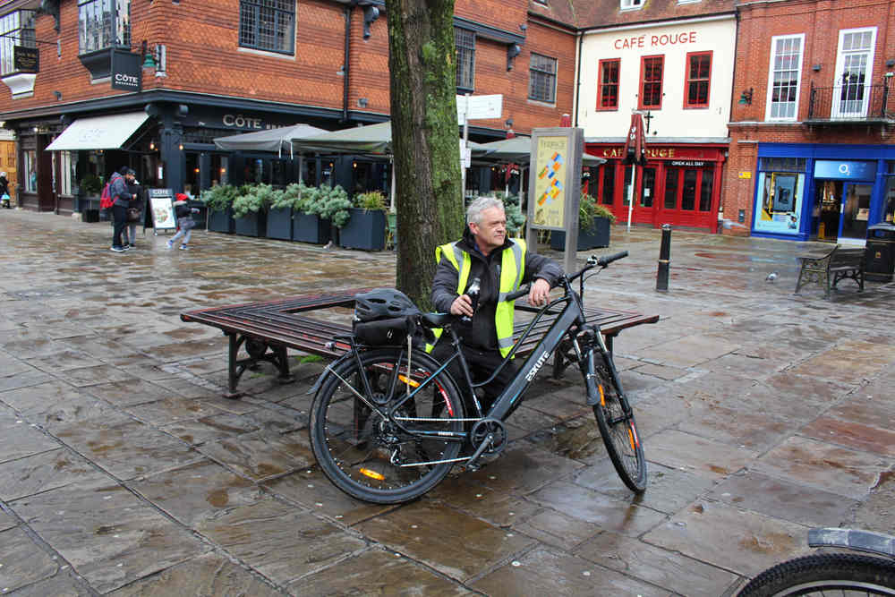 An old man sits on a bench behind a e-bike and has a drink