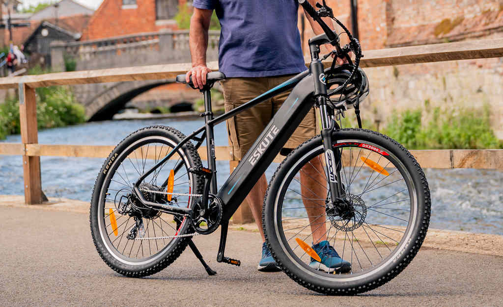  a man stands at the riverside and holds on to his electric mountain bike with derailleur gear