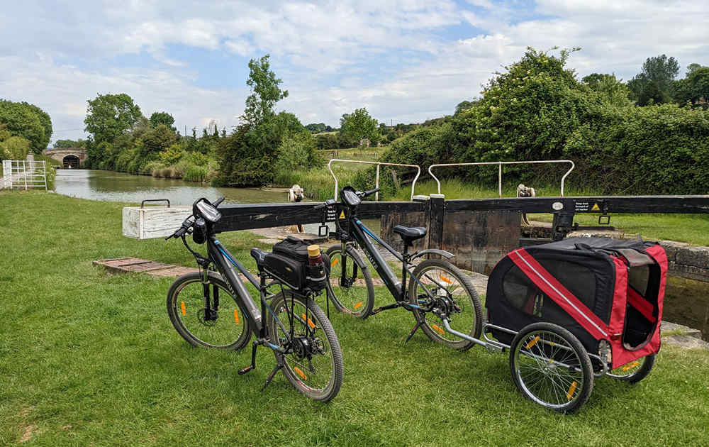 two ebikes are on the grasslands-one with bike bags on the rear rack, another one with a trailer