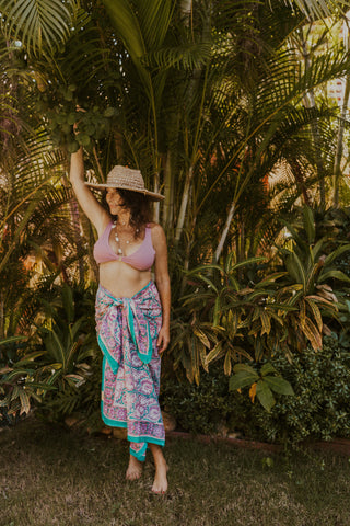 Lucia wearing our Kauai sarong in turquoise and purple floral print, standing among lush tropical plants.