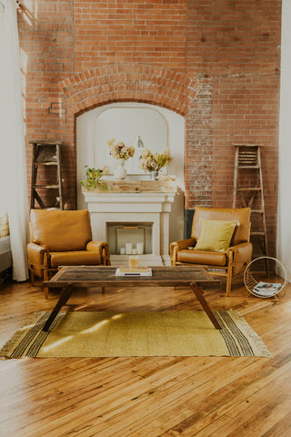 Kaiilani Turmeric Kos Rug shown under a wood coffee table in a NY style living room with exposed brick and vintage accents.