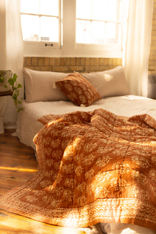 Cinnamon Kos Throw Quilt draped over the edge of a bed against a brick wall with sun shining through the large windows.
