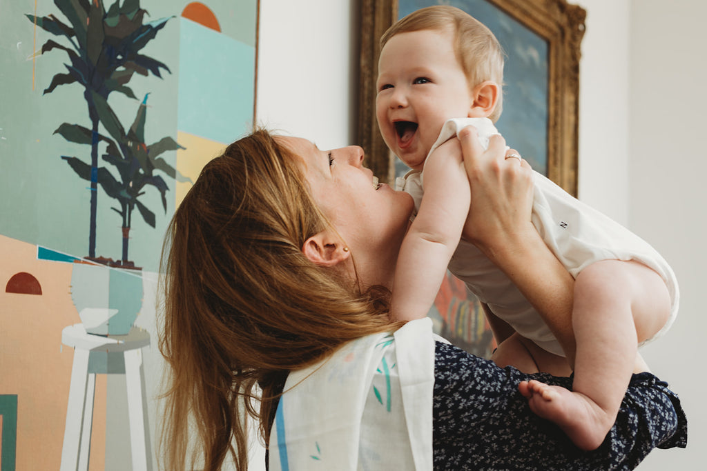 Mum holds baby boy wearing plain white baby clothes and a unique muslin on her shoulder