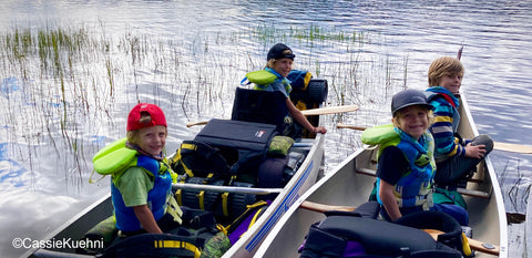 Four young boys sitting on seats and portage packs in a loaded canoe on a canoe camping trip