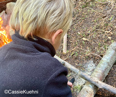 Blond child wearing fleece next to a fire cutting wood with a small saw on a canoe trip