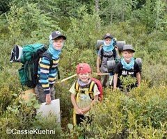 Four young boys with loaded canoe packs standing among bushes on a portage trail