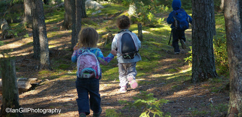 Three small children wearing backpacks running along a trail in the woods