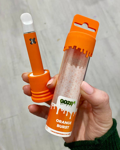 A white girl's hand is holding an Orange Burst OozeX disposable vape. The vape is connected to the base of the packaging and the top is next to it in her hand.