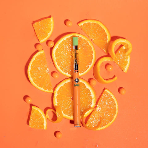 A Tropicana Punch OozeX oil cartridge is attached to an orange Ooze Slim Twist Pen that is laying on an arranged pile of orange slices.