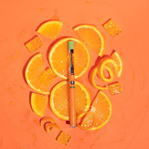 An orange Ooze Slim Twist vape pen has an OozeX oil cartridge attached with a green tip. The pen is laying on a bed of orange slices on an orange background.