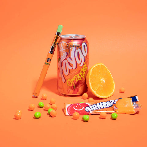 An orange Ooze Slim Twist Pen with a Tropicana Punch OozeX cartridge attached is leaning against a can of Faygo Orange Pop. There is an orange Air Head, orange and green candies, and half of an orange arranged in front.