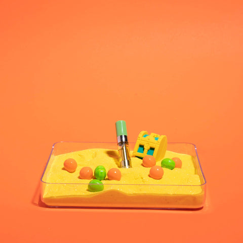 An OozeX Tropicana Punch oil cartridge is standing upright in a square dish of yellow sand with green and orange candies surrounding it.