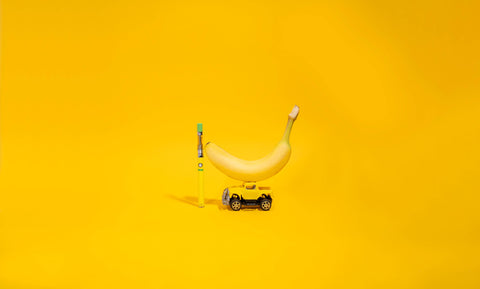 A banana is resting on a yellow toy truck next to a Sour Banana Sherber OozeX oil cartridge attached to a yellow Ooze Twist Slim Pen against a bright yellow background