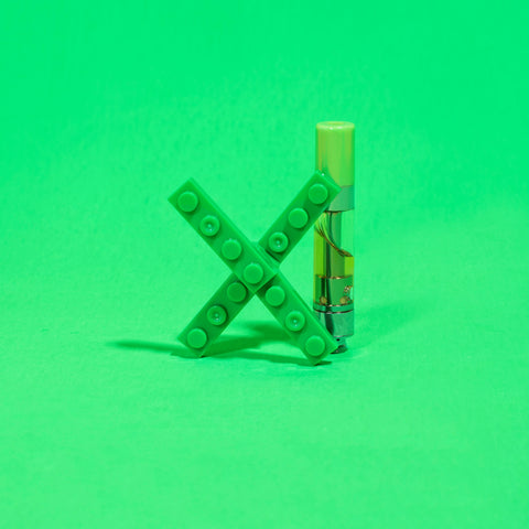 An OozeX 510 thread oil cartridge with a green mouthpiece is standing upright right behind a green X made out of skinny legos.