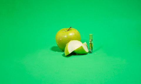 A slice of apple is next to a full granny smith apple, which is sitting next to a Sour Banana Sherbert OozeX oil cartridge against a green background.