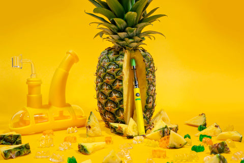 An OozeX Pineapple Trainwreck cart is attached to a yellow Ooze pen that is standing upright against a pineapple with pineapple pieces scattered in front and a yellow Ooze Steamboat in the background.
