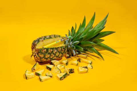 A yellow Ooze Twist Slim Pen with a Pineapple Trainwreck OozeX cartridge is laying in the crevice of a pineapple with a slice taken out and pieces scattered around.
