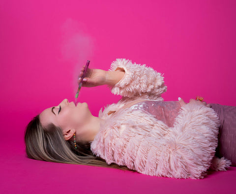 A brunette white woman wearing all pink is laying down while smoking an OozeX Strawberry Haze cartridge on a pink Ooze pen. She is exhaling smoke.