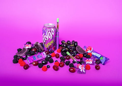 An OozeX Grape Inferno indica oil cartridge is attached to a purple Ooze Slim Twist Pen and is next to a can of grape Faygo and a pile of other grape candy