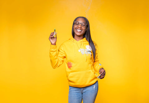 A young black girl is smiling and wearing a yellow sweatshirt and jeans is holding the yellow Ooze pen with a Sour Banana Sherbert OozeX cartridge attached.