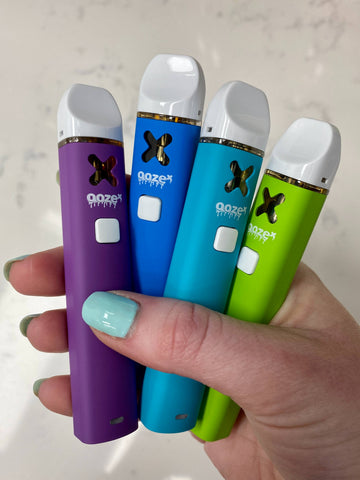 A white girl's hand with light blue nails is holding 4 OozeX HHC disposable vapes in her hand. From left to right, they are purple, dark blue, light blue, and green.