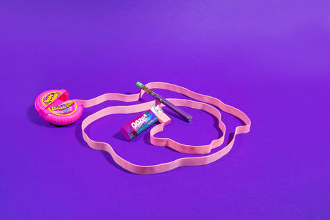 A Bubba Gum OozeX oil cartridge is attached to an Ooze pen and is laying on an unraveled roll of bubble gum tape against a bright purple background.