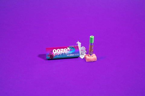A Bubba Gum OozeX 510 thread oil cartridge is standing upright on top of a piece of pink bubble gum. The bubble gum packagin is next to it and has the OozeX logo.