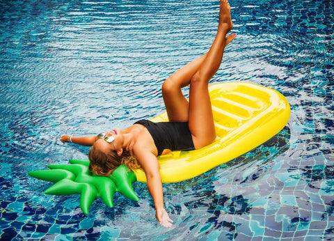 A tan woman in a strapless one-piece bathing suit is laying in a pool on a pineapple shaped float with her legs in the air and her arms hanging in the water.