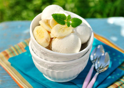 A bowl of banana sherbert with slices of bananas on top is on an outdoor table with two spoons next to the bowl.